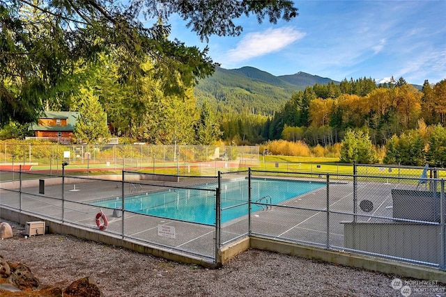 view of swimming pool with a tennis court, fence, a mountain view, and a view of trees