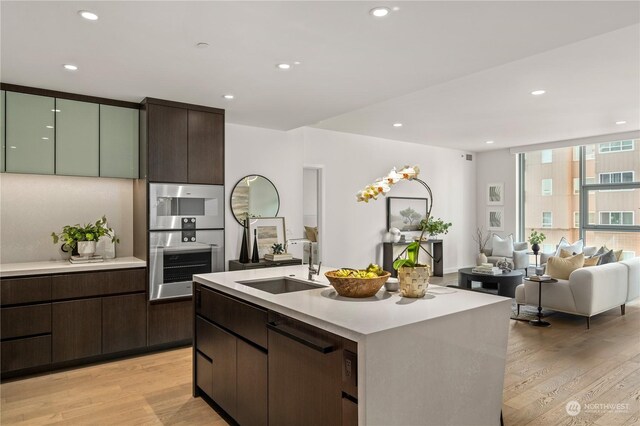 kitchen featuring light hardwood / wood-style floors, dark brown cabinetry, sink, stainless steel double oven, and an island with sink