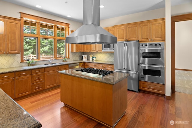 kitchen featuring island exhaust hood, appliances with stainless steel finishes, a sink, dark stone countertops, and wood finished floors