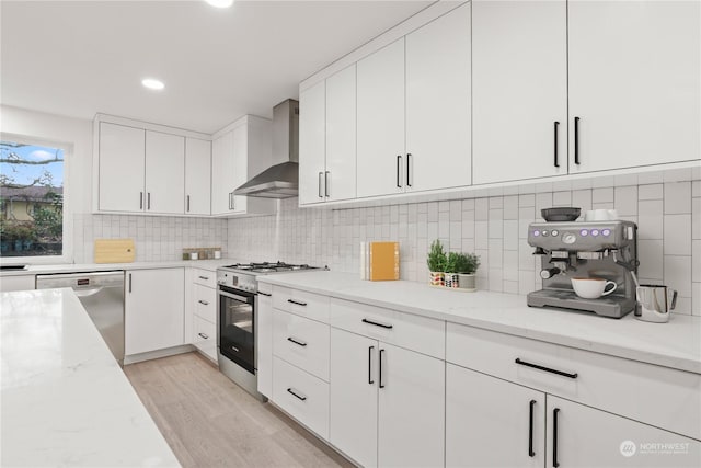 kitchen with white cabinets, stainless steel appliances, light stone countertops, wall chimney range hood, and light wood-type flooring