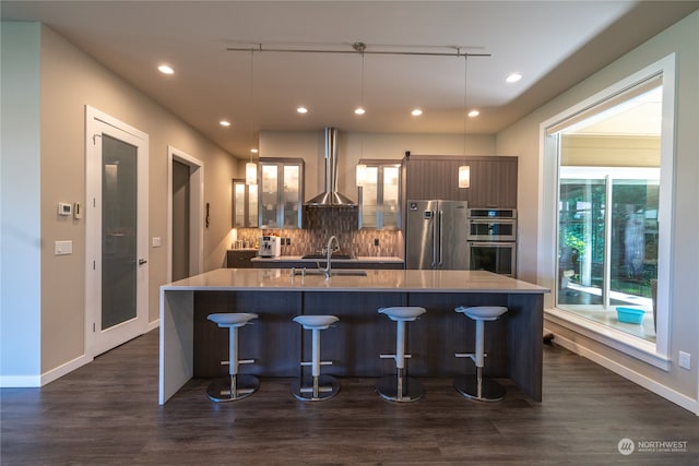 kitchen with dark hardwood / wood-style flooring, an island with sink, wall chimney exhaust hood, appliances with stainless steel finishes, and pendant lighting