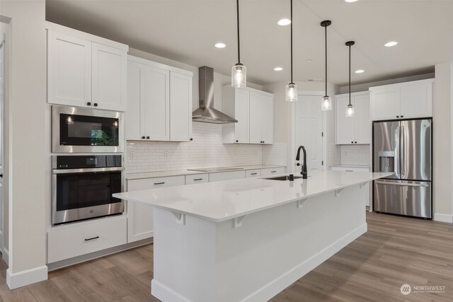 kitchen featuring stainless steel appliances, white cabinets, light countertops, wall chimney exhaust hood, and an island with sink