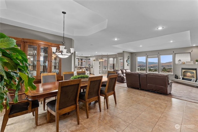 dining area with a raised ceiling, a brick fireplace, light tile patterned floors, and a notable chandelier