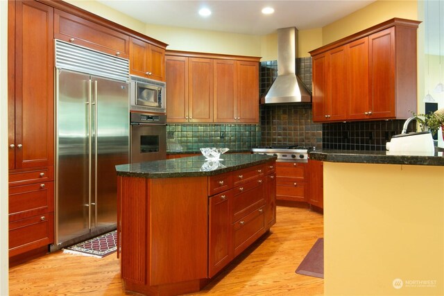 kitchen featuring a center island, wall chimney range hood, built in appliances, dark stone countertops, and light hardwood / wood-style floors