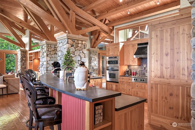 kitchen featuring a kitchen island with sink, vaulted ceiling with beams, stainless steel appliances, wooden ceiling, and wall chimney exhaust hood