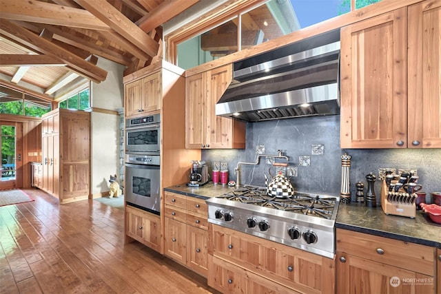 kitchen with extractor fan, appliances with stainless steel finishes, lofted ceiling with beams, wood-type flooring, and backsplash