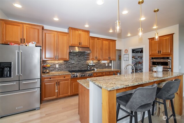 kitchen with decorative backsplash, light stone counters, an island with sink, stainless steel appliances, and light wood-type flooring