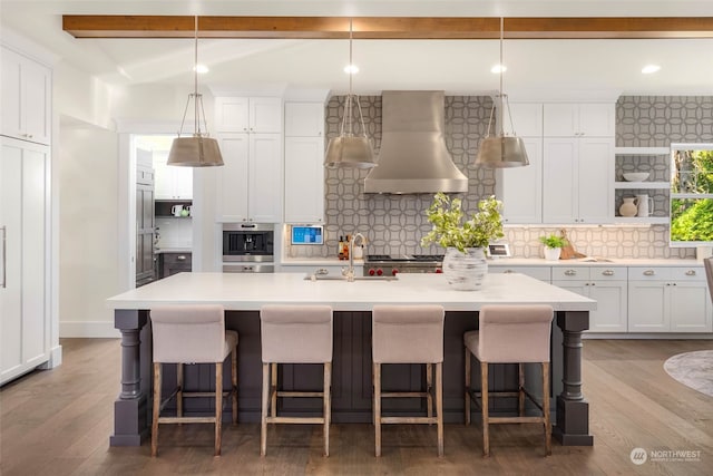 kitchen featuring wall chimney range hood, a kitchen breakfast bar, an island with sink, oven, and white cabinets