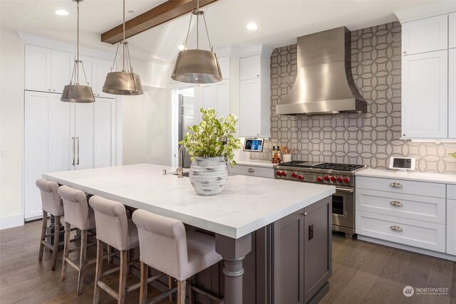 kitchen with beam ceiling, wall chimney range hood, double oven range, a kitchen island with sink, and white cabinets