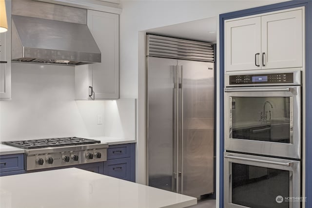 kitchen featuring wall chimney range hood, white cabinets, blue cabinets, and appliances with stainless steel finishes