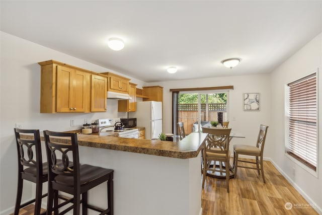 kitchen featuring white appliances, dark countertops, wood finished floors, a peninsula, and under cabinet range hood