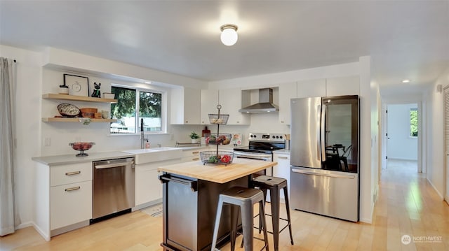 kitchen with appliances with stainless steel finishes, a healthy amount of sunlight, light wood-type flooring, a center island, and wall chimney exhaust hood