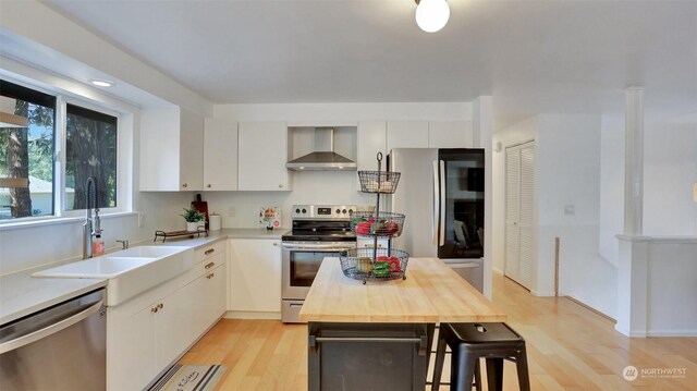 kitchen with wall chimney range hood, white cabinets, butcher block countertops, light hardwood / wood-style floors, and appliances with stainless steel finishes