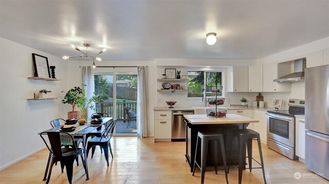 kitchen with wall chimney range hood, appliances with stainless steel finishes, white cabinets, and a healthy amount of sunlight