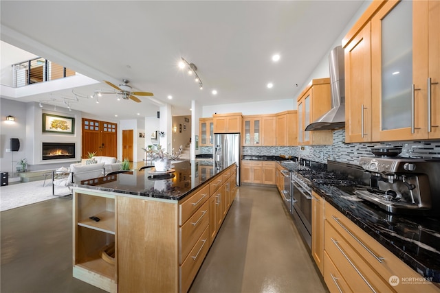 kitchen with decorative backsplash, dark stone counters, stainless steel appliances, wall chimney range hood, and a center island