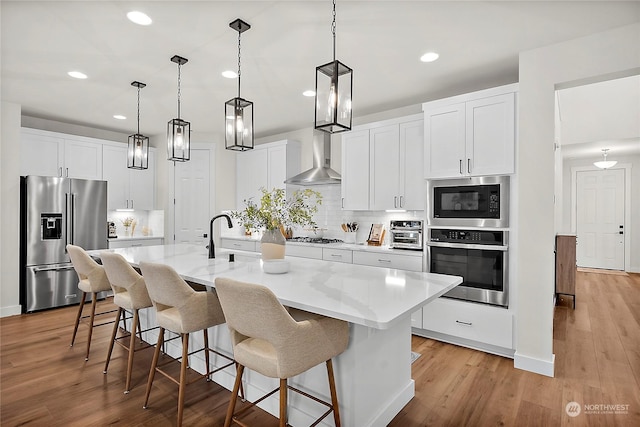 kitchen featuring appliances with stainless steel finishes, wall chimney exhaust hood, white cabinets, decorative light fixtures, and a kitchen island with sink