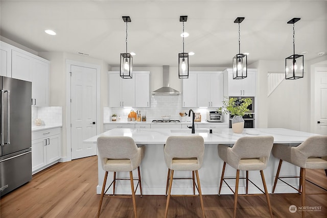 kitchen with a center island with sink, high end fridge, wall chimney exhaust hood, and white cabinets