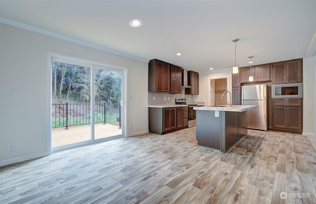 kitchen with pendant lighting, wall chimney range hood, light wood-type flooring, an island with sink, and appliances with stainless steel finishes