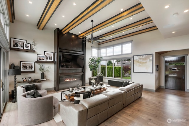 living room featuring ceiling fan, beam ceiling, a towering ceiling, a fireplace, and light wood-type flooring