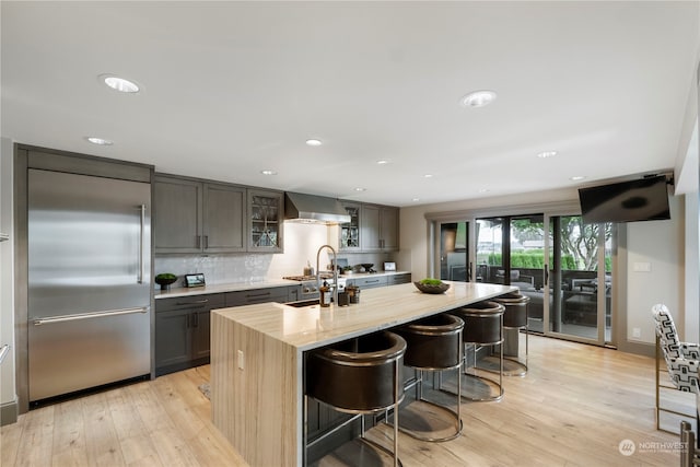 kitchen featuring a center island with sink, a kitchen breakfast bar, light wood-type flooring, stainless steel built in fridge, and wall chimney exhaust hood