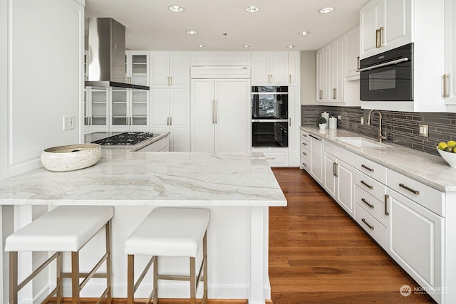 kitchen with paneled refrigerator, a sink, white cabinets, wall chimney range hood, and glass insert cabinets