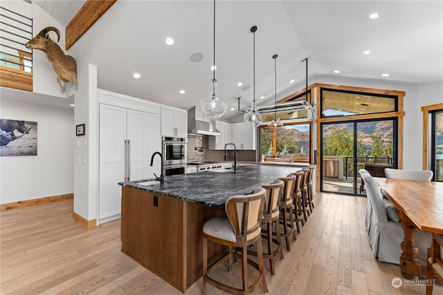 kitchen featuring a center island with sink, white cabinets, tasteful backsplash, wall chimney range hood, and dark stone counters