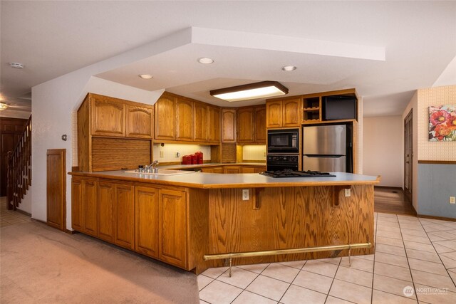 kitchen with a peninsula, a sink, light countertops, brown cabinets, and black appliances