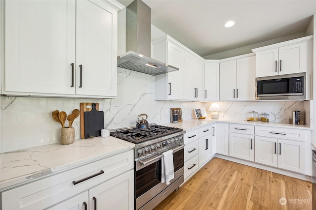 kitchen with light stone countertops, light wood finished floors, appliances with stainless steel finishes, white cabinetry, and wall chimney exhaust hood