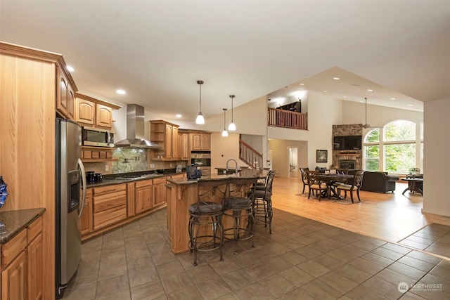 kitchen featuring wall chimney exhaust hood, open floor plan, stainless steel appliances, a kitchen bar, and a fireplace