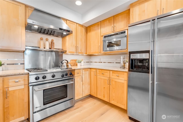 kitchen with light brown cabinets, decorative backsplash, light wood-type flooring, wall chimney exhaust hood, and high end appliances