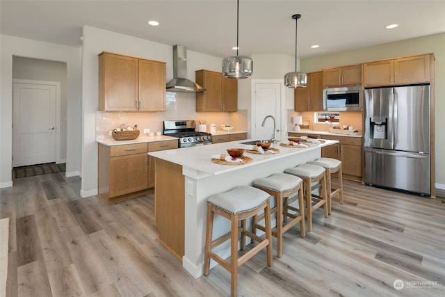 kitchen featuring appliances with stainless steel finishes, wall chimney range hood, light hardwood / wood-style floors, sink, and a center island with sink