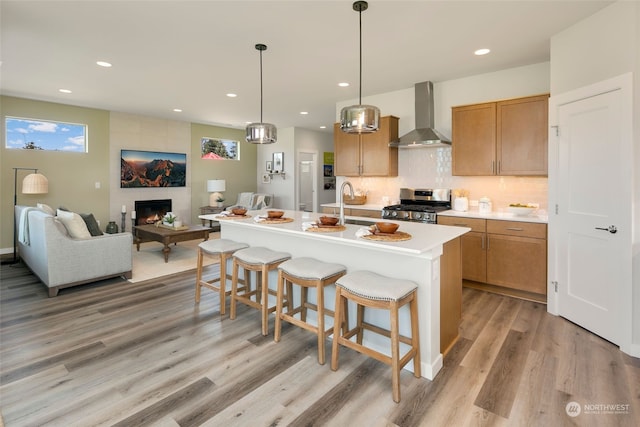 kitchen featuring a fireplace, gas range, wall chimney range hood, an island with sink, and a breakfast bar area