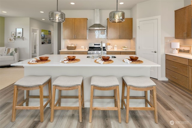 kitchen with wall chimney range hood, hanging light fixtures, stainless steel gas range, and an island with sink