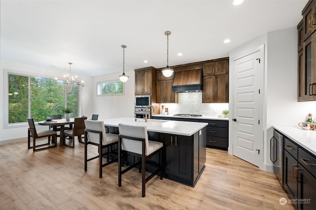 kitchen with decorative light fixtures, light wood-type flooring, appliances with stainless steel finishes, premium range hood, and a center island with sink