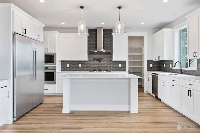 kitchen with light wood finished floors, built in appliances, light countertops, wall chimney range hood, and a sink