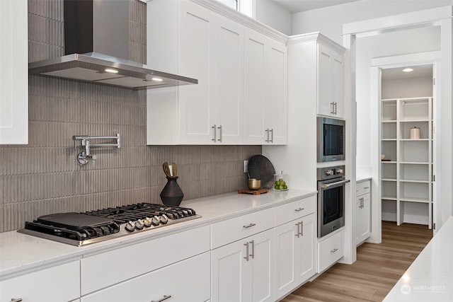 kitchen featuring appliances with stainless steel finishes, decorative backsplash, light hardwood / wood-style floors, wall chimney range hood, and white cabinets