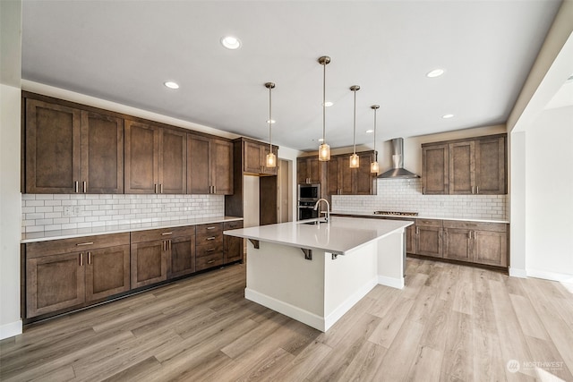 kitchen with tasteful backsplash, wall chimney range hood, light hardwood / wood-style flooring, and a center island with sink