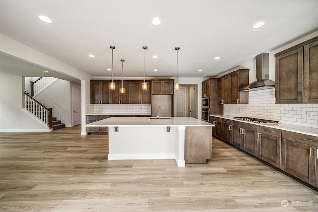 kitchen featuring wall chimney range hood, decorative backsplash, appliances with stainless steel finishes, a kitchen island with sink, and light hardwood / wood-style flooring