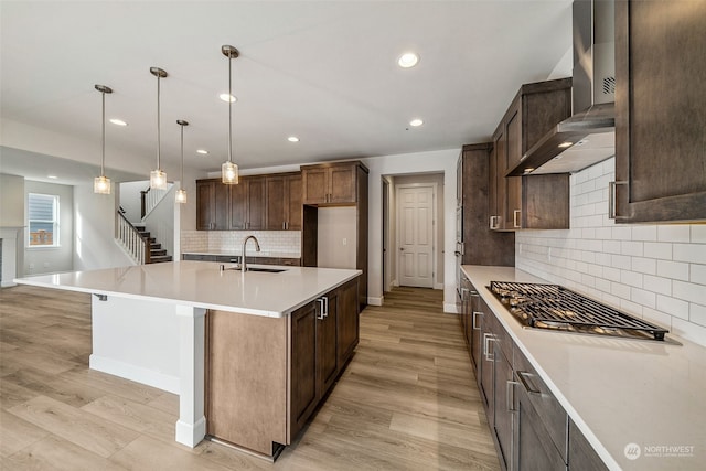 kitchen featuring a center island with sink, hanging light fixtures, light wood-type flooring, wall chimney exhaust hood, and sink