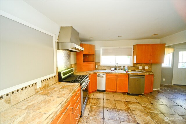 kitchen featuring decorative backsplash, tile counters, wall chimney exhaust hood, stainless steel appliances, and sink