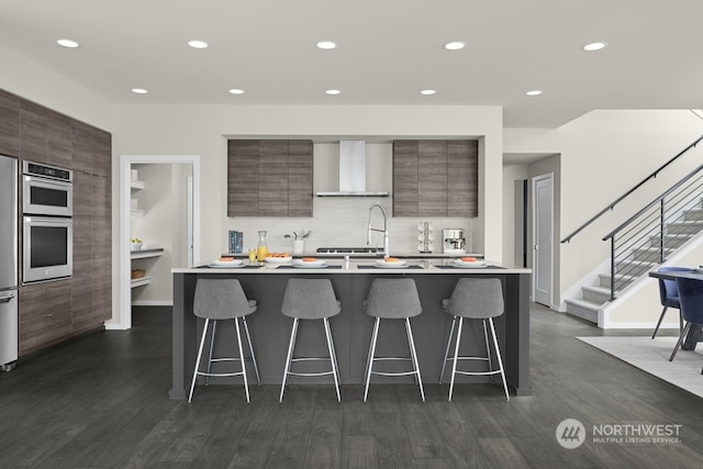 kitchen featuring backsplash, wall chimney range hood, dark wood-type flooring, a kitchen breakfast bar, and a kitchen island with sink