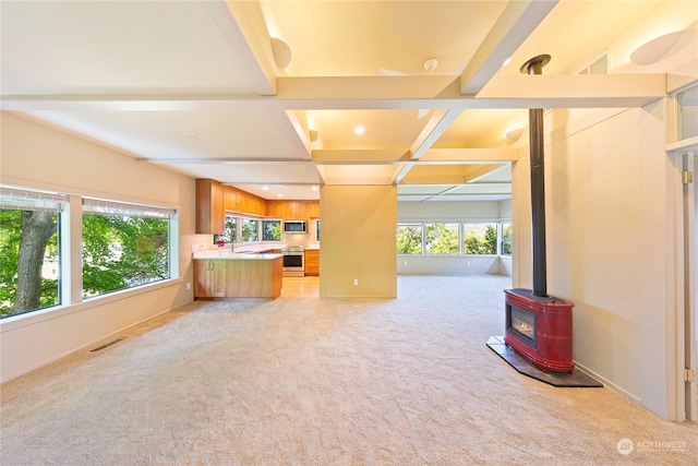 unfurnished living room featuring light colored carpet, beam ceiling, a wood stove, and a wealth of natural light