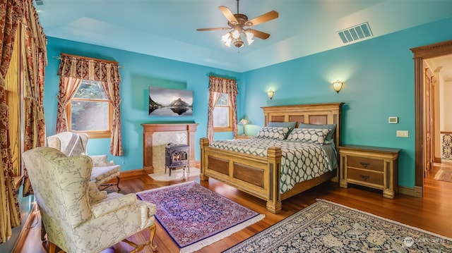 bedroom featuring multiple windows, hardwood / wood-style flooring, ceiling fan, and a wood stove