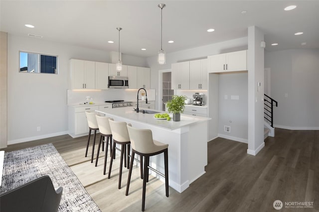 kitchen featuring wood finished floors, an island with sink, a sink, stainless steel microwave, and backsplash