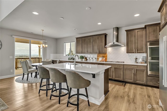 kitchen with light wood finished floors, light countertops, a kitchen island with sink, wall chimney range hood, and a kitchen bar