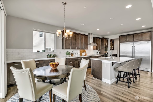 kitchen with wall chimney exhaust hood, stainless steel appliances, light hardwood / wood-style flooring, decorative light fixtures, and a center island with sink