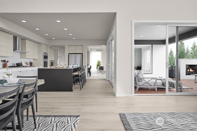 kitchen featuring light wood-style flooring, stainless steel appliances, a lit fireplace, wall chimney range hood, and modern cabinets