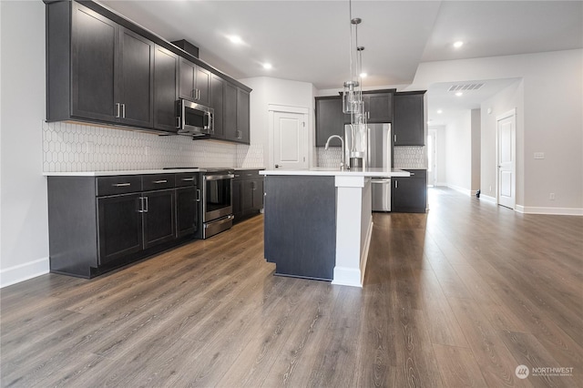 kitchen with dark hardwood / wood-style flooring, backsplash, stainless steel appliances, decorative light fixtures, and an island with sink