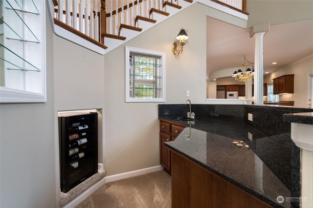 bar featuring wine cooler, dark stone counters, sink, an inviting chandelier, and decorative columns