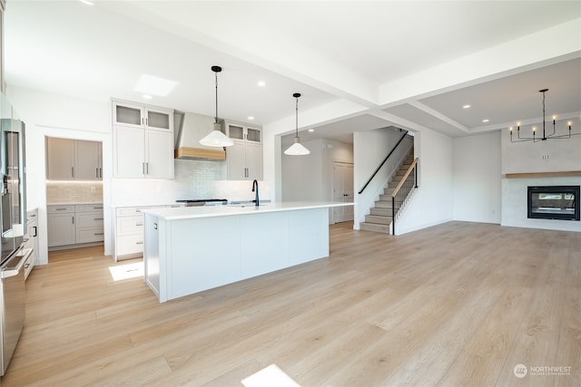 kitchen with custom exhaust hood, decorative light fixtures, an island with sink, white cabinets, and light hardwood / wood-style floors